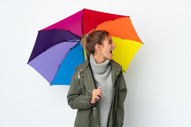 Junge Frau, die einen Regenschirm isoliert auf weißer Wand hält, die in seitlicher Position lacht