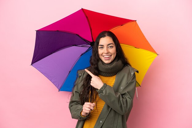 Junge Frau, die einen Regenschirm lokalisiert auf rosa Hintergrund hält, der zur Seite zeigt, um ein Produkt zu präsentieren