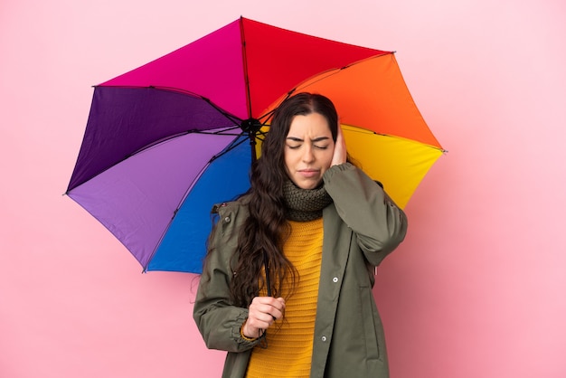 Junge Frau, die einen Regenschirm lokalisiert auf rosa Wand frustriert hält und Ohren bedeckt