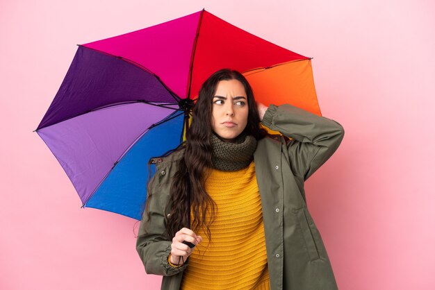 Junge Frau, die einen Regenschirm lokalisiert auf rosa Wand hält, die Zweifel hat