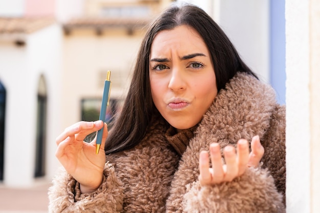 Junge Frau, die einen Stift im Freien hält und Zweifel macht, während sie die Schultern anhebt