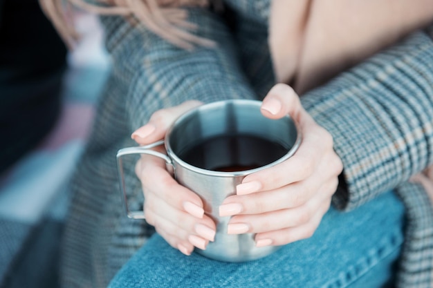 Junge Frau, die Eisenbecher mit heißem Tee hält. Nahaufnahme von weiblichen Händen, die eine Tasse heißes Getränk halten