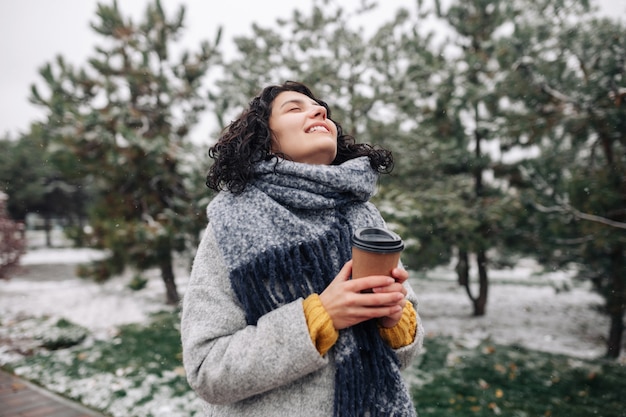 Junge Frau, die frey Modemantel und blauen Schal trägt, steht mit einem Kaffee, um an einem Winter-Schneepark zu gehen.