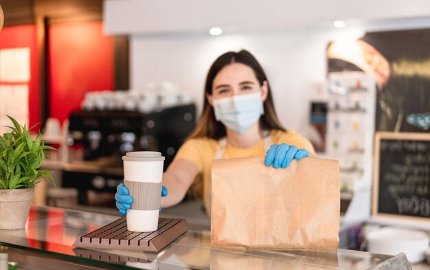 Junge Frau, die Gesichtsmaske beim Servieren Frühstück und Kaffee zum Mitnehmen in der Cafeteria trägt