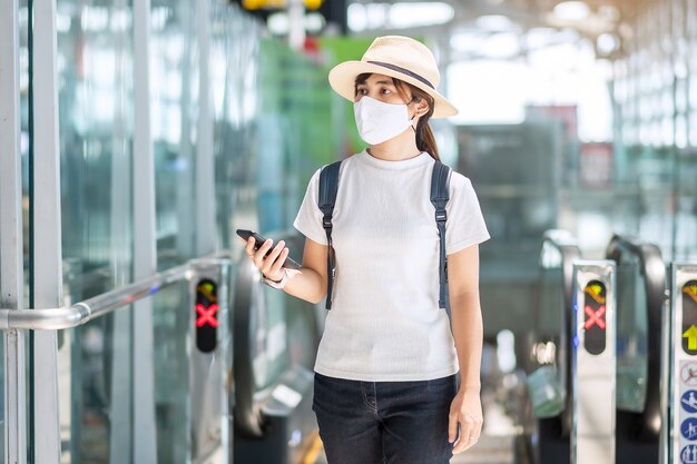 Junge Frau, die Gesichtsmaske trägt und mobiles Smartphone im Flughafen verwendet