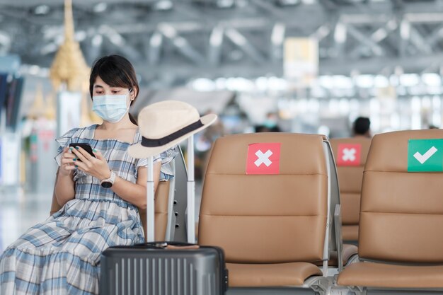 Junge Frau, die Gesichtsmaske trägt und mobiles Smartphone im Flughafen verwendet