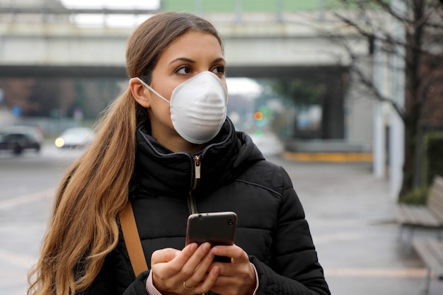Junge Frau, die Gesichtsmaske unter Verwendung des Smartphones in der Stadtstraße trägt