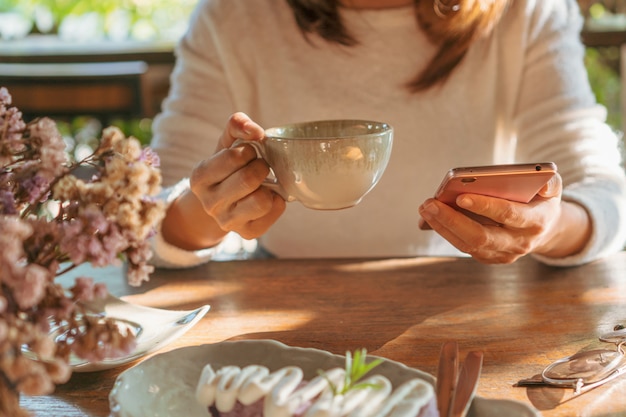 Junge Frau, die gute Nachrichten auf Handy während der Ruhe im Kaffeehaus liest, glückliche asiatische Frau, die ihr Foto auf Smartphone beim Trinken des Kaffees im Café während der Freizeit beobachtet.