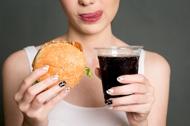 Junge Frau, die Hamburger und Kolabaum auf grauem Hintergrund isst