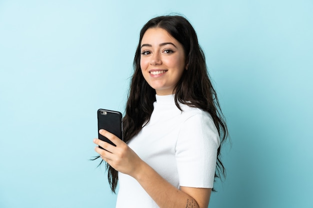 Junge Frau, die Handy lokalisiert auf blauem Hintergrund verwendet, der viel lächelt