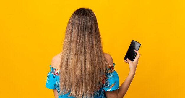 Junge Frau, die Handy über isolierte gelbe Wand in der hinteren Position verwendet