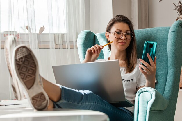 Junge Frau, die Handy und Laptop benutzt, um nach Hause zu arbeiten