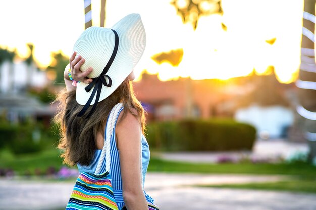 Junge Frau, die hellblaues Sommerkleid und gelben Strohhut trägt, der modische Umhängetasche hält, die draußen warmes Wetter im Sommerpark bei Sonnenuntergang genießt