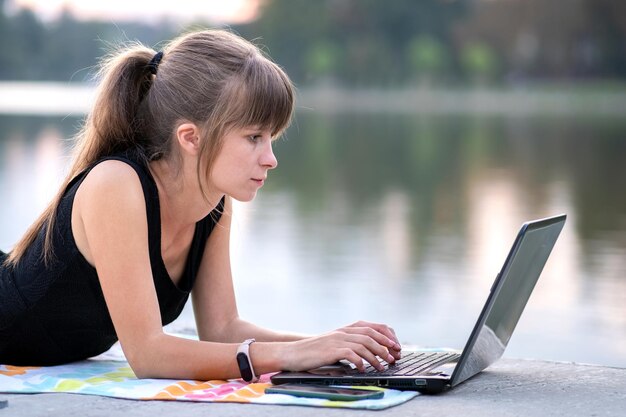 Junge Frau, die hinter einem Laptop arbeitet, der sich im Sommerpark im Freien hinlegt.