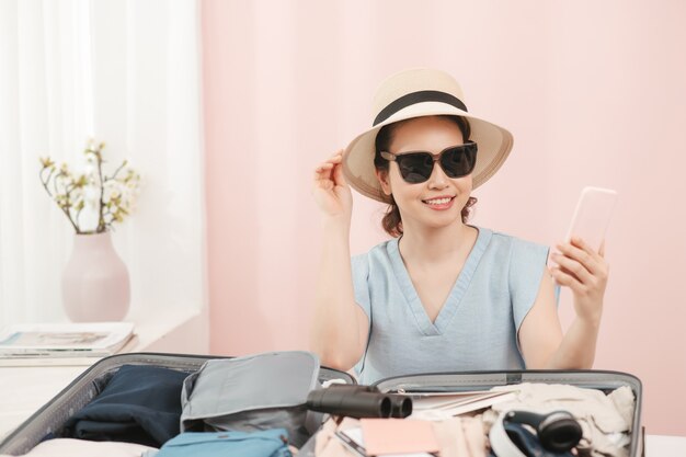 Junge Frau, die Hut wählt, der mit Kleidung und Koffer auf dem Bett sitzt. Vorbereitung auf die Sommerferien