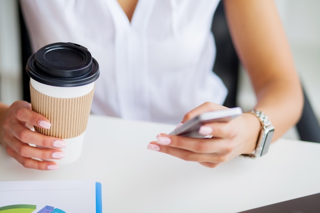 Junge Frau, die ihr Telefon verwendet und Kaffee im Büro trinkt