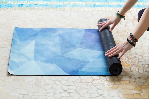 Junge Frau, die ihre blaue Yogamatte nach einer Yogaklasse auf Boden nahe einem Pool rollt
