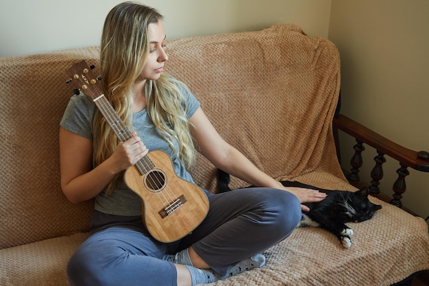 Junge Frau, die ihre Katze streichelt, während sie zu Hause auf dem Sofa sitzt und eine Ukulele in ihren Händen hält