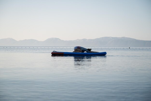 Junge Frau, die ihre morgendliche meditative Routine ausführt, sitzt in Kinderpose auf einem Sup-Board, das auf ruhigem Meerwasser schwimmt