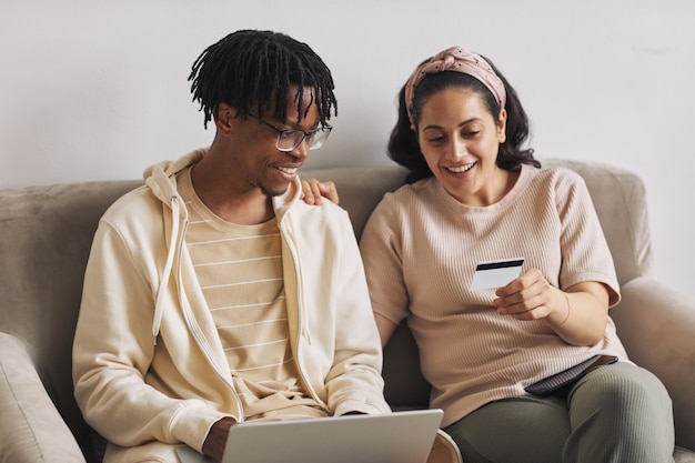 Junge Frau, die ihrem Freund die Nummer ihrer Kreditkarte zeigt, die mit Laptop online einkaufen