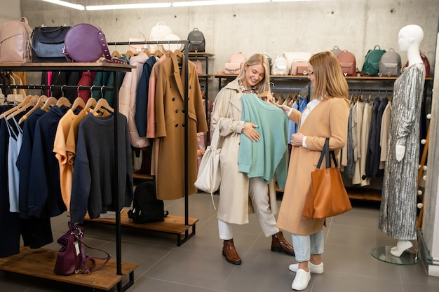 Junge Frau, die ihrem Freund hellblauen Baumwollpullover zeigt, während sie neue Kleidung in der Mall während des Einkaufens wählt