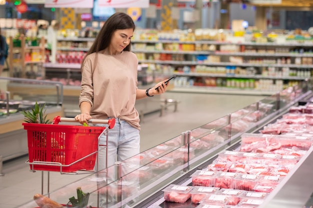 Junge Frau, die im Gang mit Einkaufswagen steht und frisches rohes Fleisch im modernen Supermarkt wählt