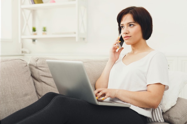 Junge Frau, die im Internet surft und zu Hause auf dem Handy spricht, auf beigefarbener Couch sitzt, Platz kopieren