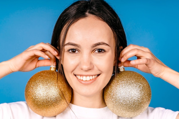 Junge Frau, die in den Händen glänzende, glänzende goldene Weihnachtskugeln blaue Farbwand hält