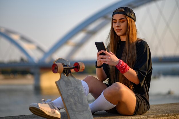 Junge Frau, die in der Nähe des Flusses sitzt und nach der Fahrt mit dem Longboard Smartphone benutzt