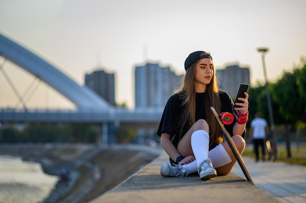 Junge Frau, die in der Nähe des Flusses sitzt und nach der Fahrt mit dem Longboard Smartphone benutzt