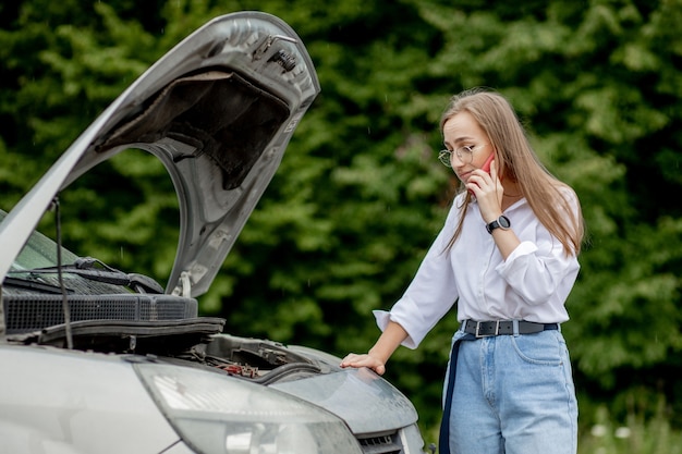 Junge Frau, die in der Nähe eines kaputten Autos mit aufgeklappter Motorhaube steht und Probleme mit ihrem Fahrzeug hat