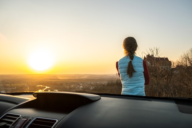 Junge Frau, die in der Nähe ihres Autos steht und die warme Aussicht auf den Sonnenuntergang genießt. Mädchenreisender, der sich auf die Motorhaube des Fahrzeugs stützt und den Abendhorizont betrachtet.