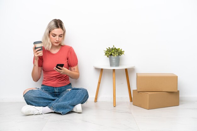 Junge Frau, die in ein neues Zuhause zwischen Kisten zieht, die auf weißem Hintergrund isoliert sind und Kaffee zum Mitnehmen und ein Handy halten