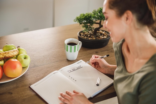 Junge Frau, die in ein Notizbuch schreibt und eine Aufgabenliste erstellt, während sie den Morgenkaffee in ihrer Küche zu Hause genießt.