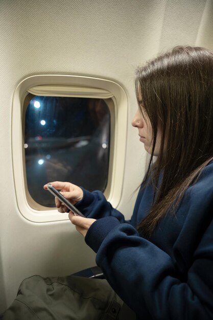 Junge Frau, die in einem Flugzeug sitzt, während sie aus dem Fenster schaut und ihr Smartphone-Reisekonzept nutzt