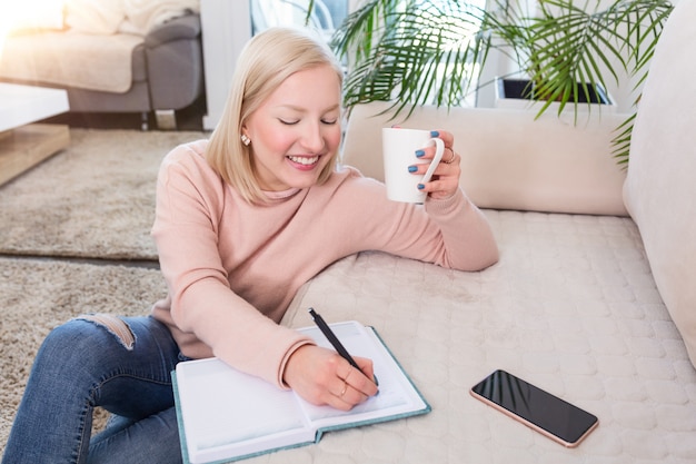 Junge Frau, die Kaffee studiert und trinkt, fleißige junge Frau, die zu Hause arbeitet und auf dem Boden im Wohnzimmer mit einer Klasse Notizen in Ordnern sitzt, die für Universität studieren