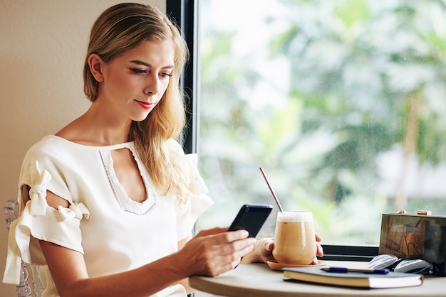 Junge Frau, die kaltes Kaffeegetränk genießt und Testnachrichten oder Artikel auf Smartphonebildschirm liest