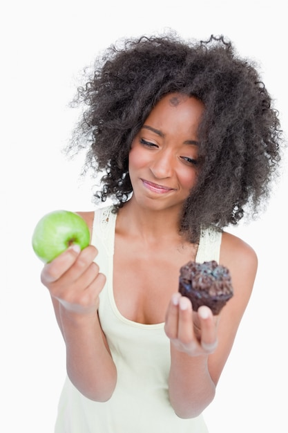 Junge Frau, die kaum zwischen einem Muffin und einem Apfel zögert