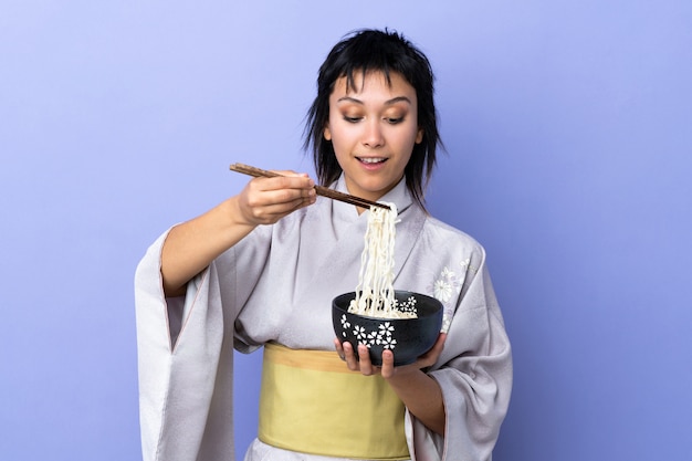 Junge Frau, die Kimono über isoliertem Blau hält, das eine Schüssel Nudeln mit Essstäbchen hält