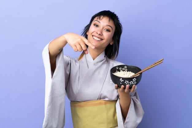 Junge Frau, die Kimono über isolierten blauen Punktfinger auf Sie mit einem selbstbewussten Ausdruck trägt, während Sie eine Schüssel Nudeln mit Stäbchen halten