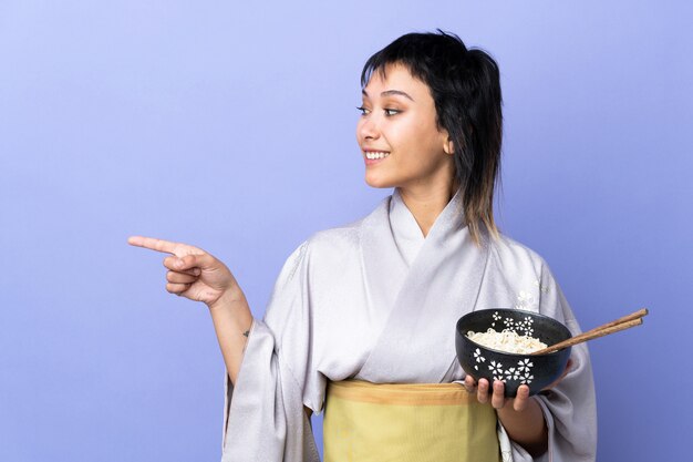 Junge frau, die kimono über isolierter blauer wand trägt, die zur seite zeigt, um ein produkt zu präsentieren, während eine schüssel nudeln mit stäbchen hält