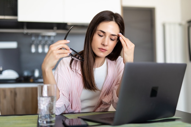 Junge Frau, die Kopfschmerzen hat, während sie am Laptop arbeitet, der an ihrem Küchentisch zu Hause sitzt.