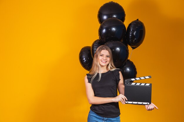 Junge Frau, die lacht, die klassische schwarze Filmherstellungsklappe hält und mit schwarzen Luftballons feiert, die über gelbem Hintergrund isoliert sind. Geburtstagsfeier, Menschen Emotionen Konzept.