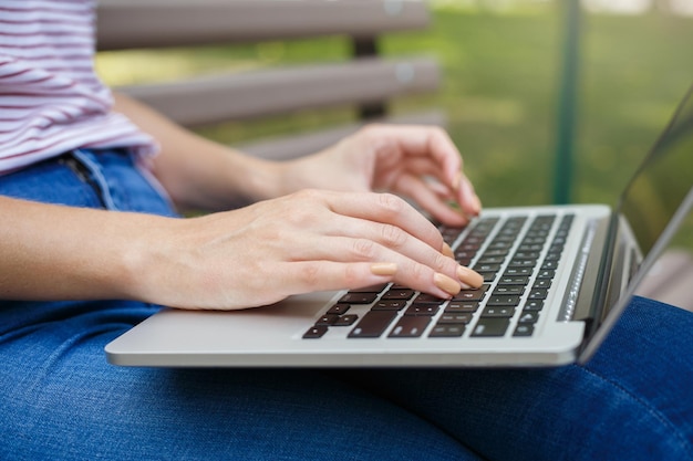 Junge Frau, die Laptop auf dem Knie hält und tippt, draußen auf der Bank sitzend. Weibliche Hand. Zuschneiden, Nahaufnahme