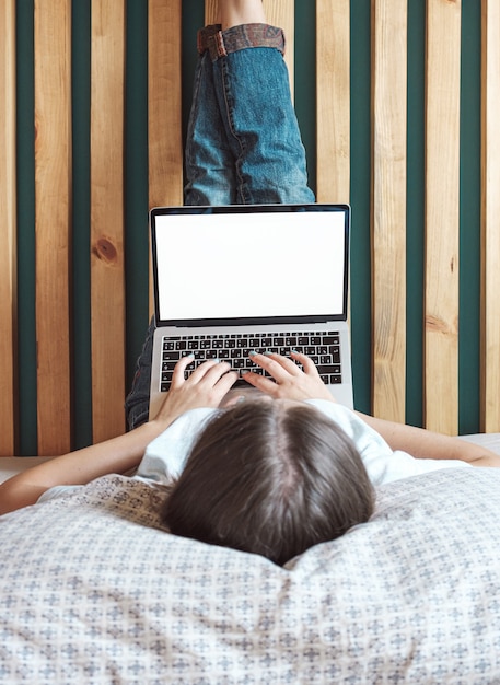 Junge Frau, die leeren Bildschirm des Laptops verwendet, um zu arbeiten, auf dem Bett im Schlafzimmer liegend, Beine hoch. Flexible Arbeitszeiten und Remote-Arbeit.