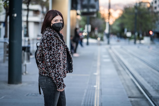 Junge Frau, die mit der schwarzen Maske gegen das Coronavirus auf die Straßenbahn wartet