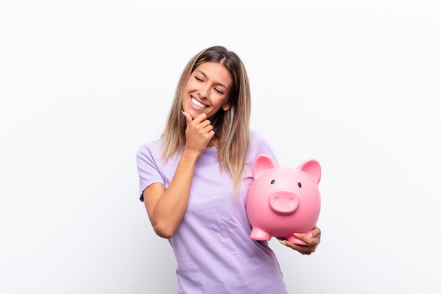 Junge Frau, die mit einem glücklichen, selbstbewussten Ausdruck mit der Hand am Kinn lächelt, sich wundert und mit einem Sparschwein zur Seite schaut.