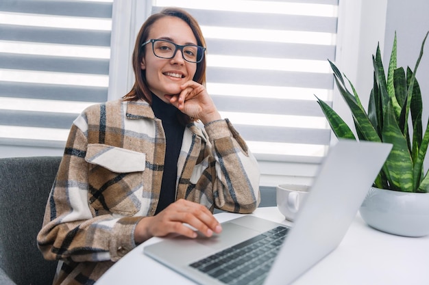 Junge Frau, die mit einem Laptop an einem Tisch sitzt und mit einem Lächeln in die Kamera schaut