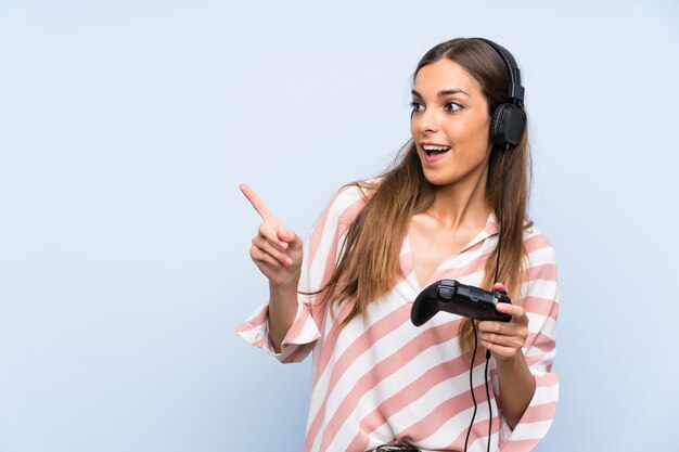 Junge Frau, die mit einem Videospielcontroller über lokalisierter blauer Wand überrascht spielt und Finger auf die Seite zeigt