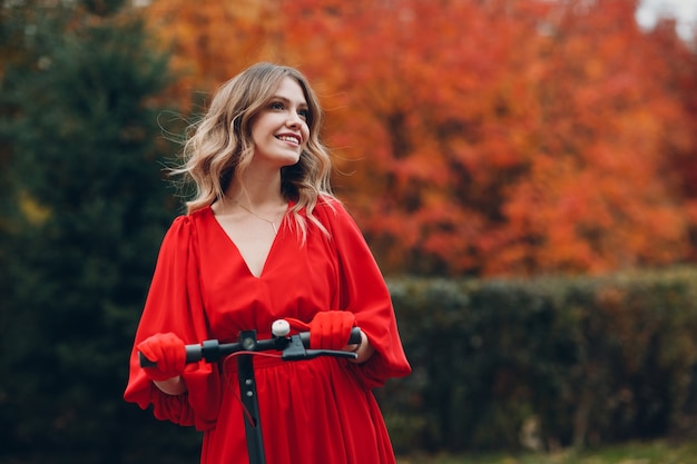 Junge Frau, die mit Elektroroller im roten Kleid und Lächeln am herbstlichen Stadtpark bleibt